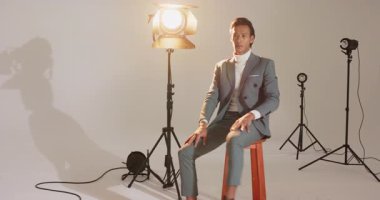 Asyalı erkek modeller anahtar ışık vardiyaları olarak stüdyodaki tabureye ayakkabı bağcıklarını ve takım elbiseyi ayarlıyorlar. Portre, moda, minimalizm, profesyonel, stil, zarafet, çağdaş