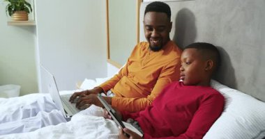 Afro-Amerikan babası oğlunu yatak odasında tabletle yatarken görmüş. Aile, bakım, konfor, sıcaklık, bağ, rahatlama, teknoloji.