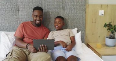 African American father tilting tablet for son leaning on bed, learning through app and smiling. Family, bonding, education, technology, warmth, comfort, interaction