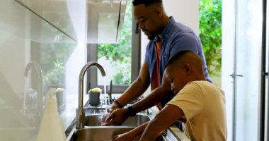 Afro-Amerikalı baba ve oğul mutfak lavabosunda bulaşık yıkıyorlar. Baba, cam yapıştırıcıları yıkıyor. Aile içi, bağ, takım çalışması, yetiştirme, yaşam tarzı, aile hayatı, ev işleri.