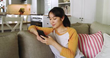 Asyalı kadın evdeki gri koltukta akıllı telefonu kaydırıyor, kameraya bakıyor ve içten bir şekilde gülümsüyor. Rahat, rahat, rahat, yaşam tarzı, iç mekan, modern, rahat