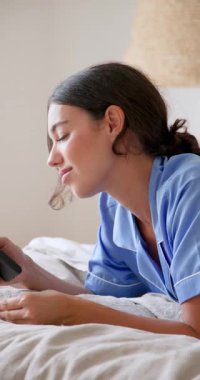 Dikey video: Pijamalı bir kadın yatakta kredi kartıyla online alışveriş yaparken gülümsüyor. ecommerce, online satın alma, yatak odası, işlem, yaşam tarzı, teknoloji