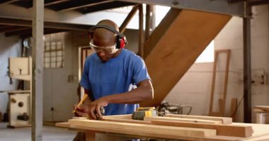 Afro-Amerikalı adam kulak koruyucularını takıyor, tahta parçasını bant ile ölçüyor, yapboz ile kesiyor. Zanaatkar, atölye, güvenlik, hassasiyet, ahşap işçiliği, marangozluk, aletler