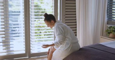 Bornozlu kadın yatakta oturmuş krema kavanozu karıştırıyor ve bacağındaki nemlendirici cilde losyon sürüyor. Cilt bakımı, bencillik, rahatlama, refah, doğal huzur, minimalizm.