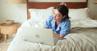 Evde pijamalı bir kadın yatakta dizüstü bilgisayar kullanıyor. Odaklanmış ve rahat görünüyor. Teknoloji, eğlence, ev ofisi, uzaktan çalışma, yaşam tarzı, çoklu görev