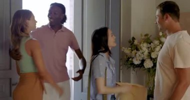 Male host opening door, welcoming diverse friends in entryway, taking hat, gesturing toward table. Friendship, hospitality, gathering, socializing, indoors, warm, casual