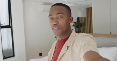 Yatak odasında Afrika kökenli Amerikalı bir adam elinde kamerayla izleyicileri selamlıyor ve el sallıyor. Vlogger, içerik oluşturucu, selfie, gündelik, modern, yaşam tarzı, iç mekan