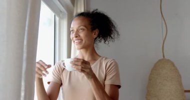 Pencere kenarında kahve içen, gülümseyen ve dışarı bakan bir kadın. Yaşam tarzı, rahatlama, tefekkür, boş zaman, sabah, şehir