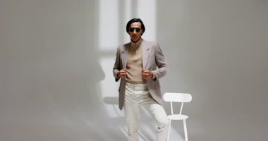 Asyalı erkek model stüdyoya giriyor, elinde siyah güneş gözlüğü, ceketi ayarlıyor, fotoshop için poz veriyor. Moda, güven, portre, minimalist, zarafet, profesyonellik, tarz