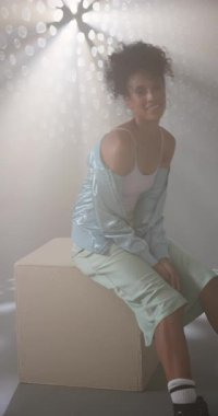 Dikey video: Şık kıyafetli genç bir kadın kutuda oturuyor, dramatik ışıklandırma altında saçlarını düzeltiyor. Moda, tarz, moda, portre, poz, cazibe