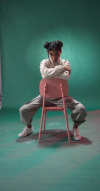 Dikey video: Genç bir kadın pembe sandalyede oturuyor, gündelik kıyafetler giyiyor, düşünceli bir şekilde aşağıya bakıyor. Moda, tefekkür, kadınlık, yalnızlık, rahatlama, yaşam tarzı.