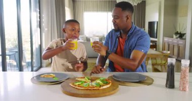 African American father and son in kitchen after boy biting pizza father clinking juice son eating. Family, bonding, domestic, nutrition, togetherness, lifestyle, warmth