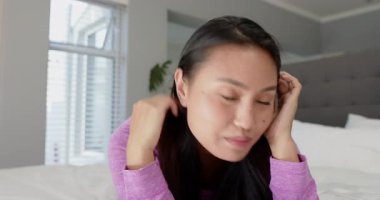 Yetişkin bir Asyalı kadın soldan giriyor, yatağa yerleşiyor, gülümsüyor, kamerayı ayarlıyor. Rahatlama, konfor, ev, yaşam tarzı, memnuniyet, doğal, samimi