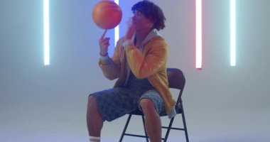 Erkek, turuncu basketbol topunu kaldırıyor, parmaklarıyla dönüyor. Neon stüdyo ışıklarında sandalyeyi kontrol ediyor. Atletik, eğlence, modern, canlı, performans, kentsel, dinamik