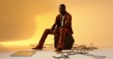 Stüdyoda Afro-Amerikan bir adam hoparlörde oturmuş gömleğinin düğmelerini açıyor ve kolsuz bluzunu çıkarıyor. Modern, moda, portre, stüdyo, günlük, tarz, güven