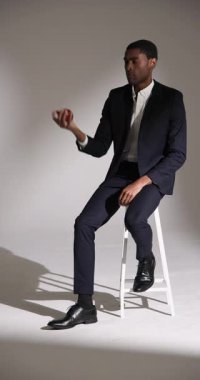 Dikey video: Takım elbiseli manken taburede oturmuş, kendinden emin görünüyor. stil, güven, zarafet, iş dünyası, modern, moda