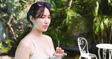 Asyalı bir kadın havuz kenarında güneşli gökyüzünün altında serinliyor. Tumbler gülerken mor içkisini yudumluyor. Lüks, rahatlık, rahatlama, canlı, açık hava, yaşam tarzı, huzur.