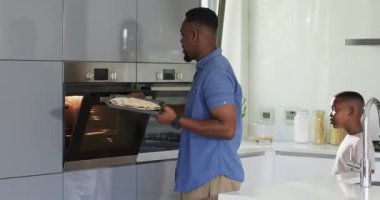 African American father and son chopping avocado, placing pizza in oven, seasoning sauce on island. Family, cooking, kitchen, bonding, preparation, modern, educational