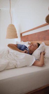 Dikey video: Kadın evinde yumuşak beyaz çarşaflarla rahat bir yatakta uyuyor. Dinlenme, rahatlama, rahatlama, uyku, sükunet, yatak.