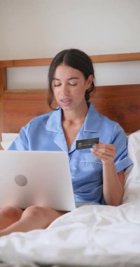 Dikey video: Mavi pijamalı kadın yatakta dizüstü bilgisayar kullanıyor, gülümsüyor ve el çırpıyor. Teknoloji, eğlence, rahatlama, yatak odası, mutluluk, ev