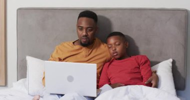 Afro-Amerikalı baba oğlunu yatakta uyurken izliyor, laptopu kapatıyor, yorganın altına sokuyor. Aile, yakınlaşma, uyku zamanı, rahatlık, şefkat, şefkat, yakınlık.