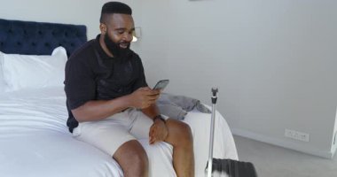Afro-Amerikalı bir adam açık renk yatak odasında, elinde siyah bavulun yanındaki akıllı telefonu tutuyor. Rahatlama, cep telefonu, bavul, minimalist, rahat, yaşam tarzı, günlük