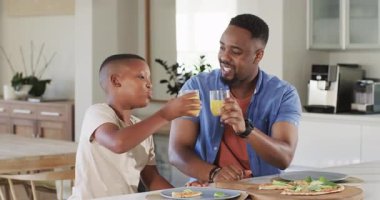 Afrika kökenli Amerikalı baba ve oğul pizza topluyor, meyve suyu dolduruyor, mutfak adasında kadeh tokuşturuyorlar. Aile, bağlılık, birliktelik, aşçılık, modern, sağlıklı, neşeli