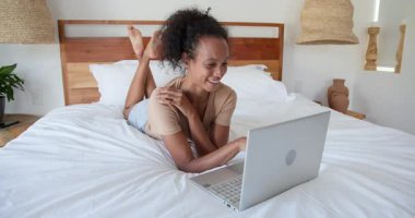 Yatakta dizüstü bilgisayar kullanan, rahat yatak odasında çalışan gülümseyen bir kadın. Uzaktan çalışma, ev ofisi, teknoloji, konfor, rahatlama, serbest çalışma