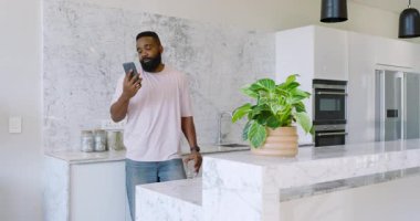 Afro-Amerikalı bir adam mutfakta tezgaha yaslanmış, elinde akıllı telefon ve cam saksı bitkileri tutuyordu. Modern, rahatlama, mutfak, yaşam tarzı, teknoloji, parlaklık, dekor