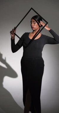 Dikey video: Stüdyoda geometrik çerçeveli zarif siyah elbiseyle poz veren model. Moda, zarafet, tarz, portre, yüksek moda, minimalizm