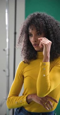 Dikey video: Sarı kazaklı kadın kıvırcık saçlı güvenli bir şekilde stüdyoda poz veriyor. Moda, özgüven, portre, bukleler, modellik, canlı