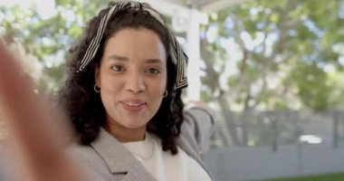 Blazer ceketli bir kadın telefon kamerasını engelliyor. Veranda avlusunu işaret eden seyirciyi selamlıyor. Gündelik, dostane, açık hava, yaşam tarzı, canlı, modern, doğal