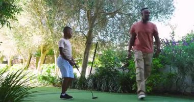 Afrikalı Amerikalı baba oğlunun vuruşuna rehberlik ediyor ve çocuğun ev yeşili üzerinde pratik yapmasını izliyor. Kopya alanı. Açık hava, aile, akıl hocalığı, öğretici, boş zaman, eğlence, büyüme