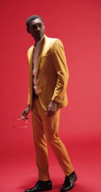 Dikey video: Sarı takım elbiseli şık bir adam güneş gözlüğünü ayarlıyor, stüdyoda güven içinde poz veriyor. Moda, tarz, erkek giyim, aksesuarlar, güven, moda