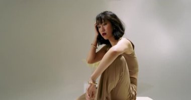 Çinli bir kadın stüdyodaki beyaz küpü indiriyor ve altın bileziği fotoşopa göre ayarlıyor. Minimalist, zarif, duruş, sofistike, dinginlik, moda, estetik