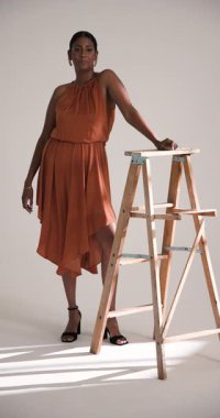 Dikey video: Şık elbiseli zarif kadın stüdyoda tahta merdivenin yanında güven içinde poz veriyor. Moda, zarafet, tarz, güzellik, güven, modellik