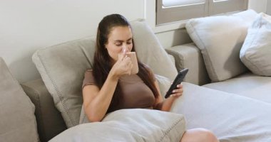 Kadın bardakları ve akıllı telefonları kaldırıyor, telefonları kaydırıyor ve evdeki kanepede yudumluyor. Rahatlık, rahatlık, rahatlık, modern, gündelik, yaşam tarzı, evcil