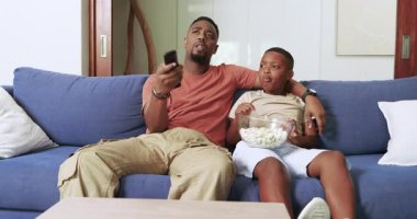 Afrika kökenli Amerikalı baba ve oğul, uzaktan kumandayla patlamış mısır yiyen mavi kanepede televizyon kanallarını tarıyor. Aile, bağ, rahatlama, rahatlama, ev, konfor, bağlantı