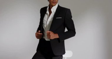 Asyalı erkek model, beyaz taburede oturuyor. Stüdyoda çekime hazırlanan siyah ceketi ayarlıyor. Profesyonel, moda, minimalist, erkeksi, kendine güvenen, gösterişli, çok yönlü