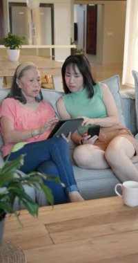 Dikey video: İki kadın kanepede tablet ve akıllı telefon kullanıyor, evde neşeli sohbetler ediyorlar. Teknoloji, iletişim, arkadaşlık, yaşam tarzı, boş zaman, cep telefonu