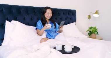 Yatağa gelen siyah tepsiyi fark eden beyaz bardaktan yudumlayan yetişkin bir kadın ve kaydırma tableti. Rahat, rahat, uyku, rahatlık, konfor, ev, yaşam tarzı.