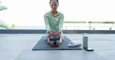 Mavi bantlı Asyalı kadın balkon minderinde havluyla gerilmek için içe doğru çekiyor. Fitness, esneklik, sağIık, rahatlama, açık hava, minimalizm, gençleşme