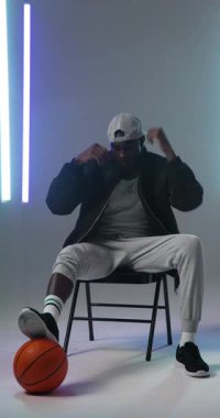 Dikey video: Basketbol tutan genç adam, sandalyede oturuyor, günlük giysi tarzında giyiniyor. Sporcu, spor, şehir, moda, eğlence, gençlik