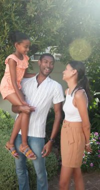 Dikey video: Anne konuşurken baba kızını kucağına alıyor, bahçede ailece vakit geçiriyor. Beraberlik, bağlılık, ebeveynlik, açık hava, mutluluk, çocukluk