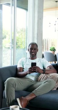 Dikey video: Evde dinlenen, koltukta oturan, akıllı telefon tutan, sohbetin tadını çıkaran bir adam. Yaşam tarzı, eğlence, teknoloji, iletişim, konfor, bağlantı