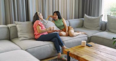 Asyalı anne ve kız evdeki kanepeye yerleşiyor, beyaz kupalara uzanıyor ve hikayelerini paylaşıyorlar. Aile, sohbet, yakınlaşma, samimiyet, rahatlama, evcil, samimi