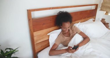 Kadın yatakta dinleniyor, akıllı telefon kullanıyor ve evde gülümsüyor. Rahatlama, yaşam tarzı, teknoloji, mutluluk, rahatlık, rahatlık