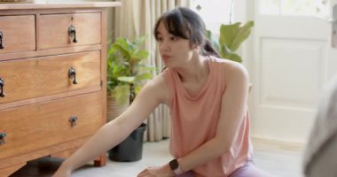 Isınmak için eve giren Asyalı kadın yoga minderinde esneme hareketleri yapıyor ve akıllı bakışlarını kontrol ediyor. Fitness, esneklik, sağIık, farkındalık, rahatlama, ev ortamı, doğal ışık