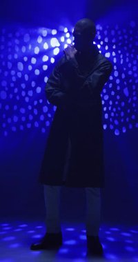 Dikey video: Stüdyoda mavi ışıkların altında uzun paltolu Afro-Amerikalı bir adam. Moda, portre, stil, modern, ışıklandırma, modellik