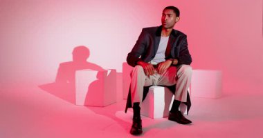 African American man entering studio, sitting on cube, checking wristwatch under pink lighting. Contemplation, professionalism, minimalism, elegance, introspection, mood, sophistication